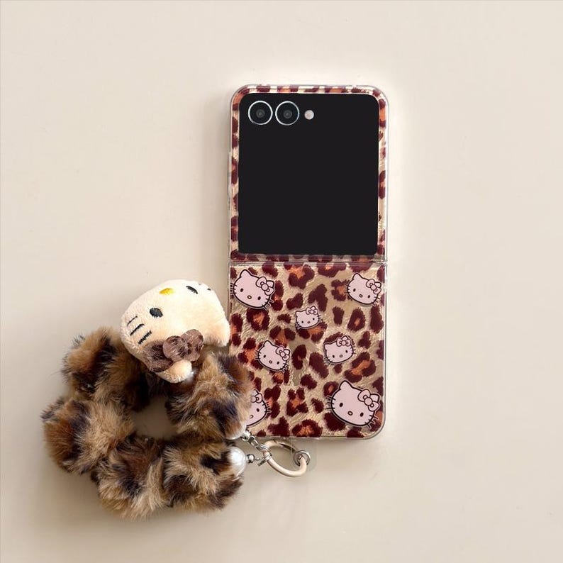 May include: A clear phone case featuring a leopard print pattern and Hello Kitty motifs. A matching fluffy scrunchie with a Hello Kitty charm is attached. The phone case is open, displaying a black screen.
