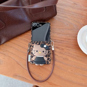 May include: A phone case featuring a Hello Kitty design with a black bow and dress, set against a leopard print background. The case has a brown strap and is next to a brown leather bag.