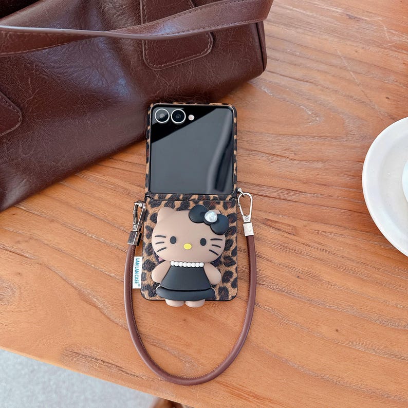 May include: A phone case featuring a Hello Kitty design with a black bow and dress, set against a leopard print background. The case has a brown strap and is next to a brown leather bag.