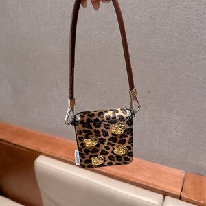 May include: A small, square case with a brown and black leopard print pattern. The case is adorned with four gold-colored crown embellishments and has a brown leather strap. The case is held by a brown leather strap with silver-colored metal clasps.