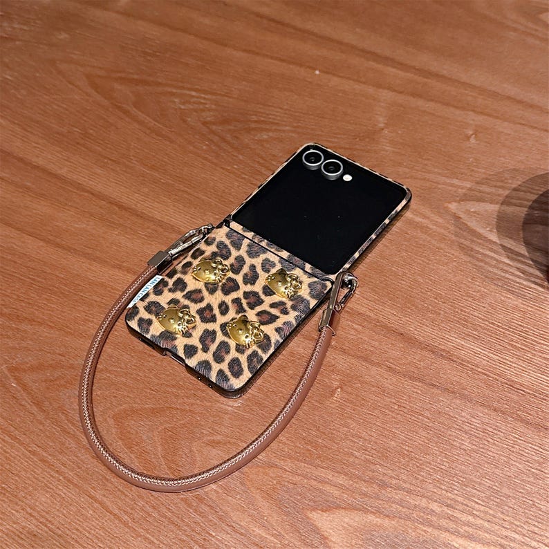 May include: A leopard print phone case with gold-colored Hello Kitty embellishments and a matching strap. The phone case is open, revealing the phone's screen and camera. The case is resting on a wooden surface.