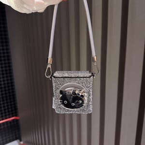 May include: A silver glittery square bag with a white strap. The bag features a black Hello Kitty face with a silver bow. The bag is held by a white strap with silver hardware.