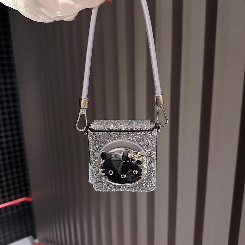 May include: A silver glittery square bag with a white strap. The bag features a black Hello Kitty face with a silver bow. The bag is held by a white strap with silver hardware.