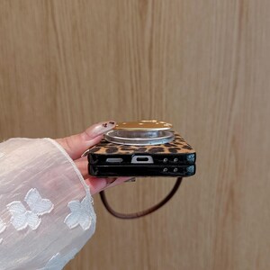 以下が含まれることがあります： ヒョウ柄の携帯電話ケースと金色のポップアップグリップ。ケースは黒い縁と茶色のストラップが付いています。ポップアップグリップは円形で、金色の金属仕上げです。ケースは人が持っています。