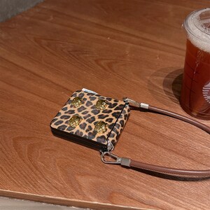 May include: A small, square leopard print wallet with gold-colored lion head studs. The wallet has a brown leather strap and a silver-colored clasp. A clear plastic cup of iced tea is in the background.