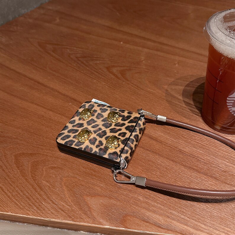May include: A small, square leopard print wallet with gold-colored lion head studs. The wallet has a brown leather strap and a silver-colored clasp. A clear plastic cup of iced tea is in the background.