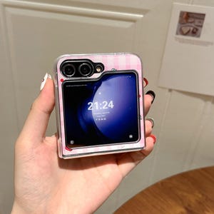 May include: A pink and white plaid phone case with a clear border, holding a phone displaying the time 21:24. The case has a camera cutout and a small strawberry design.