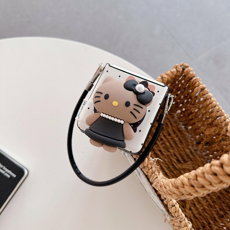 Op de afbeelding: Een wit en zwart gestippelde hoes met een 3D cartoon kat ontwerp. De kat heeft een zwarte strik, zwarte jurk en een parelketting. De hoes heeft een zwarte band en zilveren metalen accenten. De hoes ligt naast een gevlochten mand.