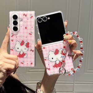 May include: Pink phone case with a Hello Kitty design. The case features a gingham pattern with cherries and a Hello Kitty graphic. A beaded wrist strap with red and clear beads is attached to the phone case.