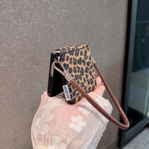 以下が含まれることがあります： ヒョウ柄の携帯電話ケースと茶色のリストストラップ。ケースは黒と茶色で、繰り返しのパターンがあります。リストストラップは茶色の革製で、シルバーの留め金が付いています。「LAN LAN CASE」の文字がケースにプリントされています。