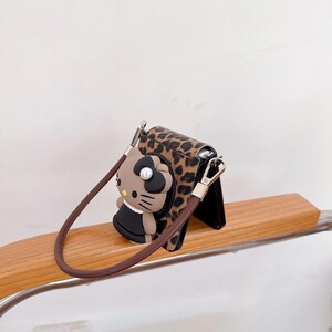 May include: A brown and black leopard print phone case with a brown strap. The case features a 3D cartoon character with a black dress and a pearl. The case is resting on a wooden surface.