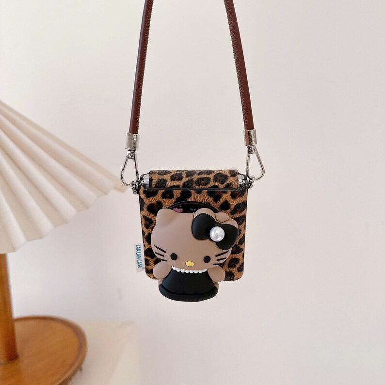 May include: A brown and black leopard print case with a brown strap, designed to hold small electronics. The case features a 3D Hello Kitty character with a black bow and pearl accent. The text "LALAN CASE" is visible on the side.