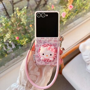 May include: A pink and white phone case featuring a Hello Kitty design. The case has a leopard print pattern and a pink strap. The phone is black with a white border. The strap has the text "LAN LAN CASE".