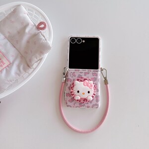 May include: A pink and white phone case featuring a Hello Kitty design. The case has a leopard print pattern and a 3D Hello Kitty figure with a pink bow. A pink lanyard is attached to the case.