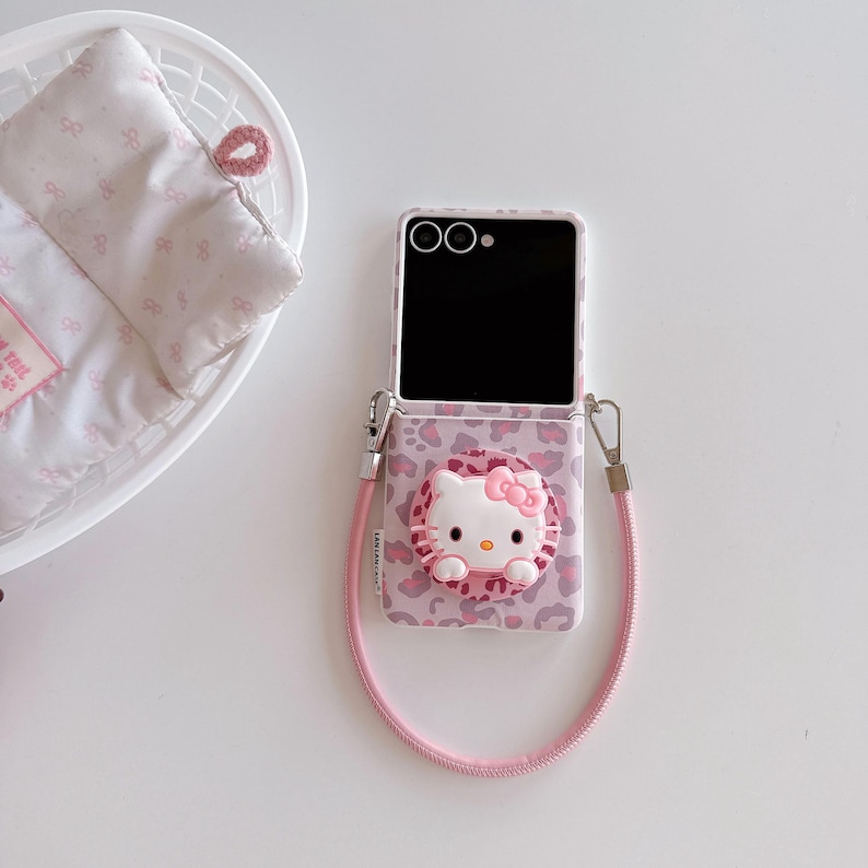 May include: A pink and white phone case featuring a Hello Kitty design. The case has a leopard print pattern and a 3D Hello Kitty figure with a pink bow. A pink lanyard is attached to the case.