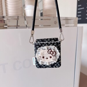 May include: A black floral print case with a white lace trim and a Hello Kitty design. The case has a black strap and silver-tone metal hardware. The case has a small white tag that reads "LAN LAN CASE".