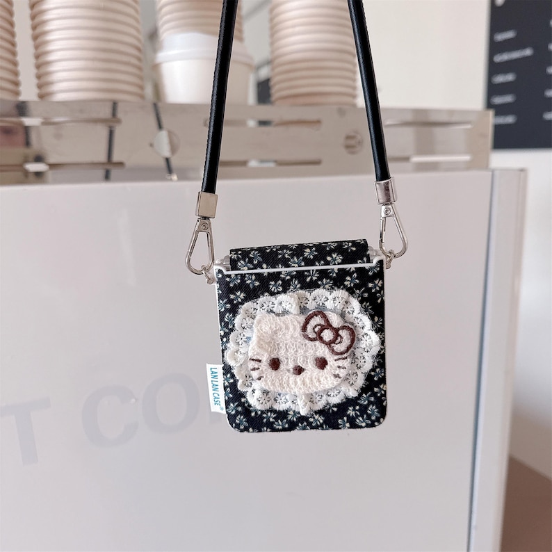 May include: A black floral print case with a white lace trim and a Hello Kitty design. The case has a black strap and silver-tone metal hardware. The case has a small white tag that reads "LAN LAN CASE".