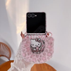 Op de afbeelding: Een roze en witte telefoonhoes met een Hello Kitty-ontwerp. De hoes heeft een roze luipaardprint en een pluizig, roze handvat. Het scherm van de telefoon is zwart.