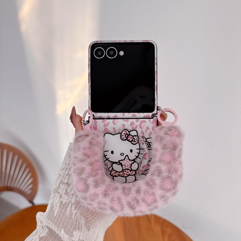 Op de afbeelding: Een roze en witte telefoonhoes met een Hello Kitty-ontwerp. De hoes heeft een roze luipaardprint en een pluizig, roze handvat. Het scherm van de telefoon is zwart.