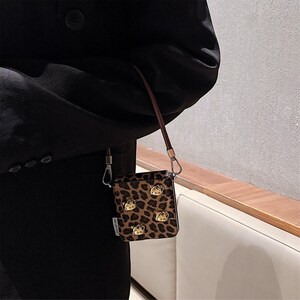 May include: A small, rectangular crossbody bag with a leopard print pattern. The bag features gold-colored embellishments and a brown strap. The bag is held by a person wearing a black blazer.