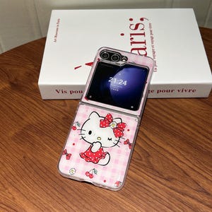 May include: A clear phone case featuring a pink gingham pattern and a cartoon image of Hello Kitty. The case is open, revealing a phone screen displaying the time 21:24. The case has a red bow and cherry accents.
