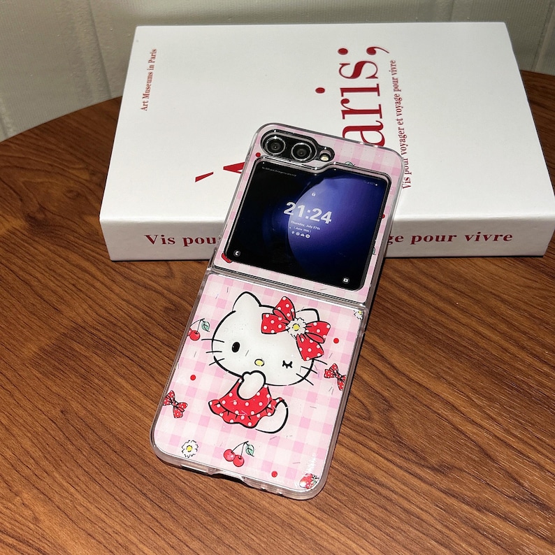 May include: A clear phone case featuring a pink gingham pattern and a cartoon image of Hello Kitty. The case is open, revealing a phone screen displaying the time 21:24. The case has a red bow and cherry accents.