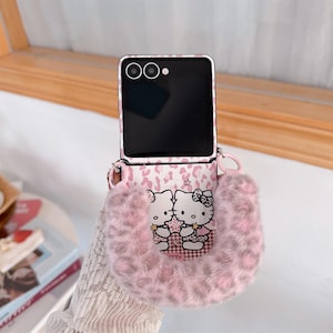 Op de afbeelding: Een roze en witte telefoonhoes met een pluizig, harig zakje. De hoes heeft een Hello Kitty-ontwerp en een roze luipaardprintpatroon. De telefoon is zwart met een zilveren frame en heeft een riem.