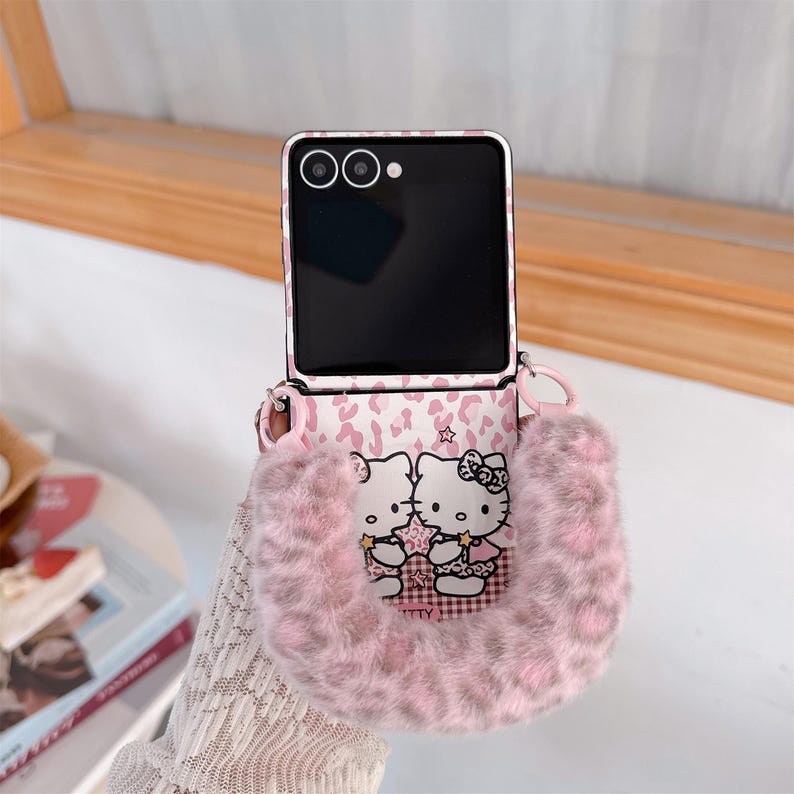 Op de afbeelding: Een roze en witte telefoonhoes met een pluizig, harig zakje. De hoes heeft een Hello Kitty-ontwerp en een roze luipaardprintpatroon. De telefoon is zwart met een zilveren frame en heeft een riem.