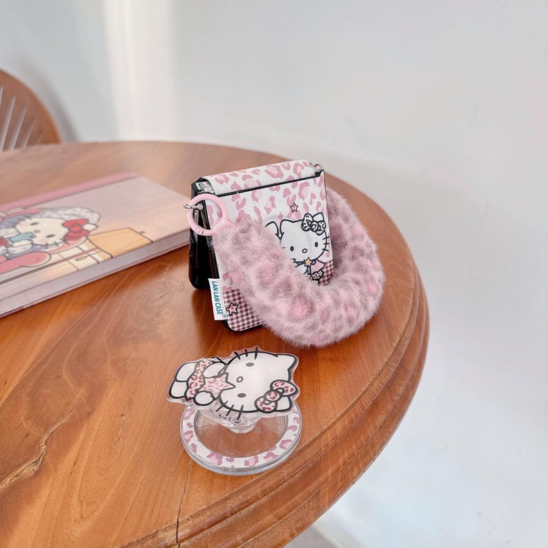 Op de afbeelding: Een roze en wit patroon hoesje met een Hello Kitty ontwerp, een bijpassende fluffy polsband en een doorzichtige telefoonhouder met een Hello Kitty afbeelding. Een notitieboek met een Hello Kitty ontwerp is ook zichtbaar.