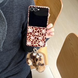 May include: A clear phone case featuring a leopard print and Hello Kitty motifs. The case is paired with a fluffy keychain in brown and tan. The phone's screen is black.