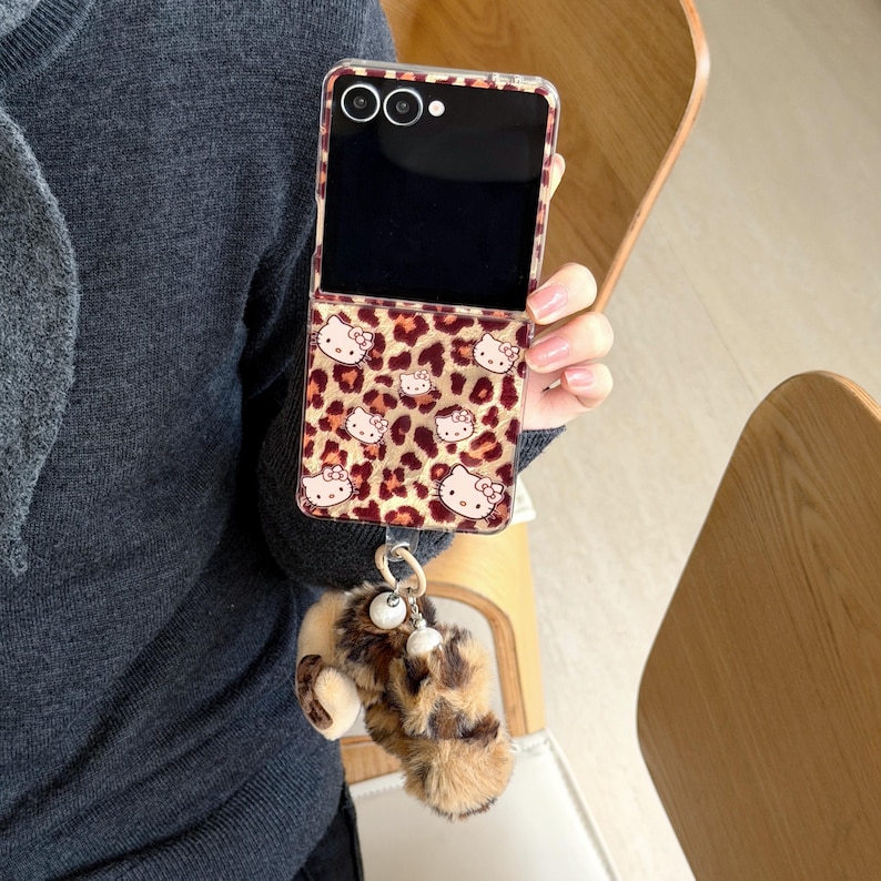 May include: A clear phone case featuring a leopard print and Hello Kitty motifs. The case is paired with a fluffy keychain in brown and tan. The phone's screen is black.