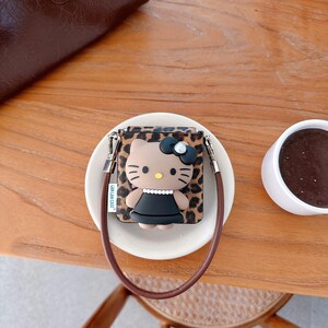 May include: A brown and tan leopard print case featuring a Hello Kitty design with a black bow and dress. The case has a brown strap and silver hardware. The case is sitting on a white plate.