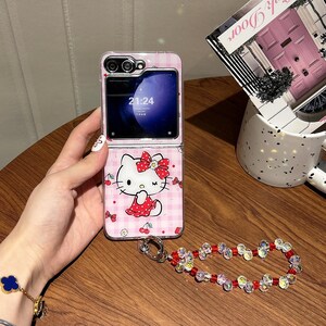 May include: A pink and white patterned phone case featuring a Hello Kitty design. The phone case is open, displaying the phone screen with the time 21:24. A decorative beaded wrist strap with red and clear beads is attached.
