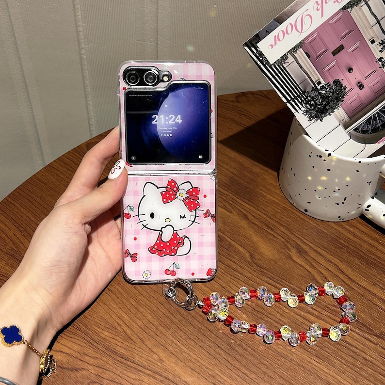May include: A pink and white patterned phone case featuring a Hello Kitty design. The phone case is open, displaying the phone screen with the time 21:24. A decorative beaded wrist strap with red and clear beads is attached.