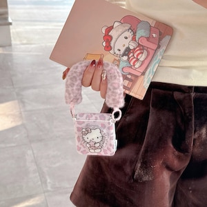 Op de afbeelding: Een roze en wit patroon AirPod-hoesje met een Hello Kitty-ontwerp. De hoes heeft een pluizig handvat en een bijpassende kaart met een Hello Kitty-illustratie. De hoes wordt vastgehouden voor een persoon die een cr&egrave;mekleurige top en bruine shorts draagt.