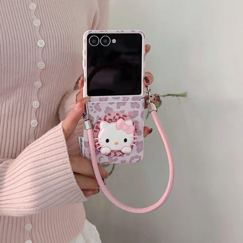 May include: A pink and white phone case with a Hello Kitty design. The case has a leopard print pattern and a pink strap. The phone is open, revealing the screen. The case is held by a person.
