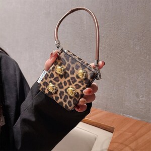 May include: A small, square handbag with a leopard print pattern and gold-colored cat head studs. The bag has a brown handle and silver-colored hardware. The text "LAI LAN CASE" is visible on the side of the bag.