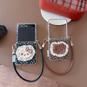 May include: Two open flip phone cases with decorative designs. One case features a white lace Hello Kitty design on a black floral background. The other case has a brown Hello Kitty design on a brown floral background. Both cases have straps.