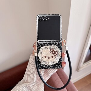 May include: A black and white floral patterned phone case featuring a Hello Kitty design. The case has a black strap and silver hardware. The Hello Kitty design is surrounded by white lace.