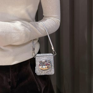 May include: A small, square, silver glitter handbag with a silver strap. The bag features a round, silver emblem with a cat design. The bag is held by a silver strap.