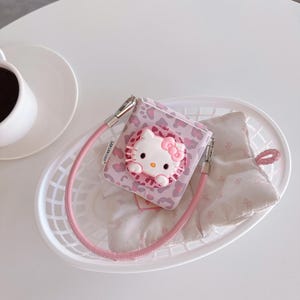 May include: A pink and white Hello Kitty-themed accessory case with a matching pink strap. The case features a 3D Hello Kitty design with a pink bow and is set against a pink leopard print background. The case is inside a white basket.