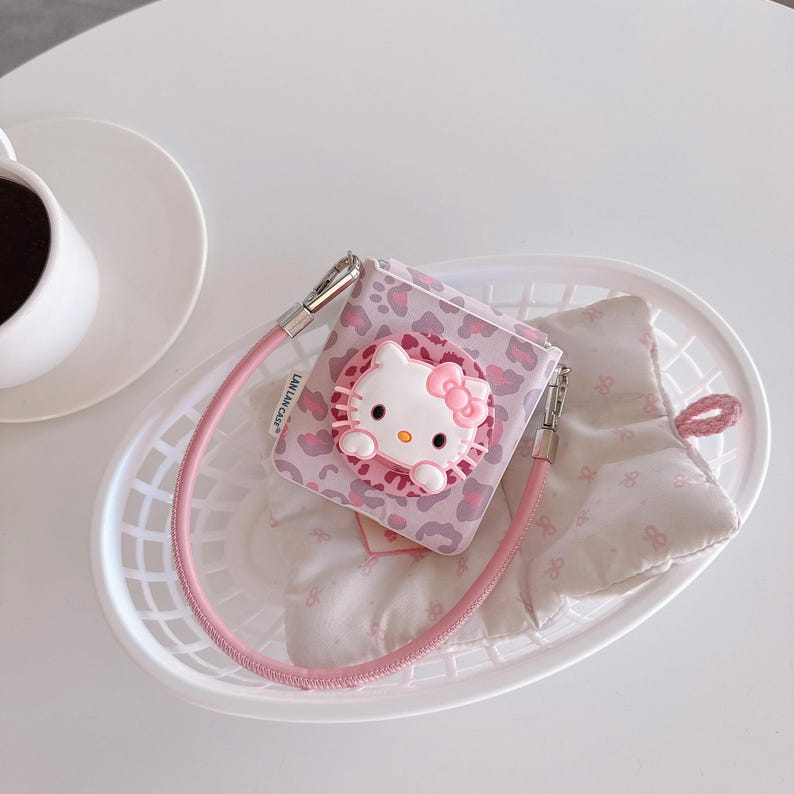 May include: A pink and white Hello Kitty-themed accessory case with a matching pink strap. The case features a 3D Hello Kitty design with a pink bow and is set against a pink leopard print background. The case is inside a white basket.