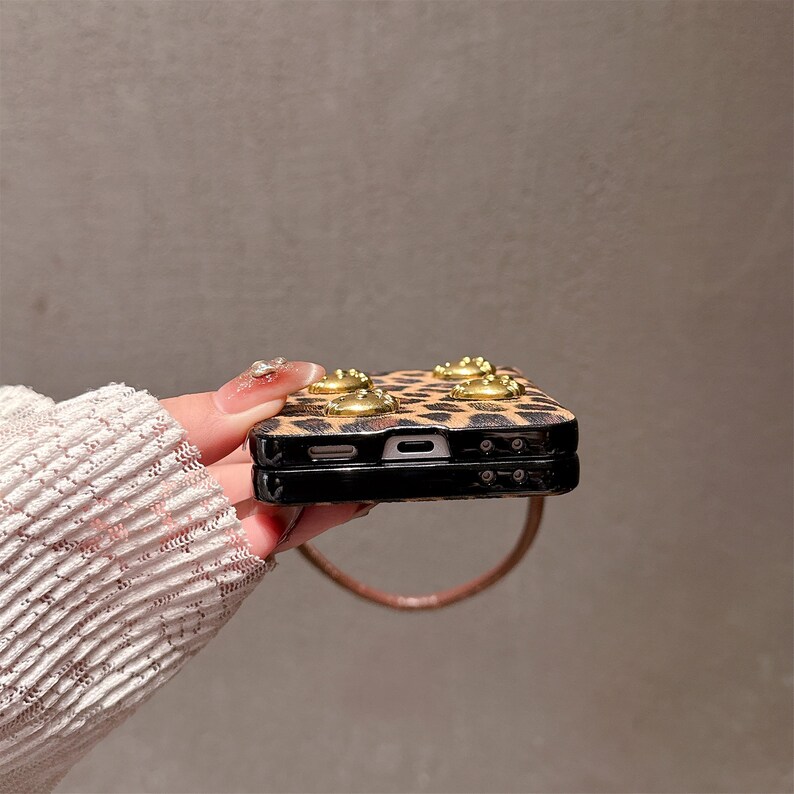 May include: A leopard print phone case with gold-colored embellishments. The case is black with a brown and black spotted pattern. The case has a brown strap attached. The phone case is a protective accessory.