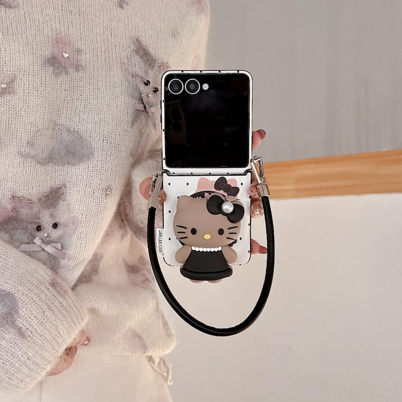 Op de afbeelding: Een wit en zwart telefoonhoesje met een cartoon kattenpersonage met een zwarte strik en jurk. De telefoonhoes heeft een zwarte band en een zwart telefoonscherm. De telefoonhoes is open.