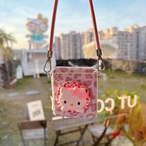 May include: Pink crossbody phone case featuring a Hello Kitty design. The case has a leopard print pattern and a 3D Hello Kitty face with a pink bow. It includes a pink strap and silver-colored metal clasps.