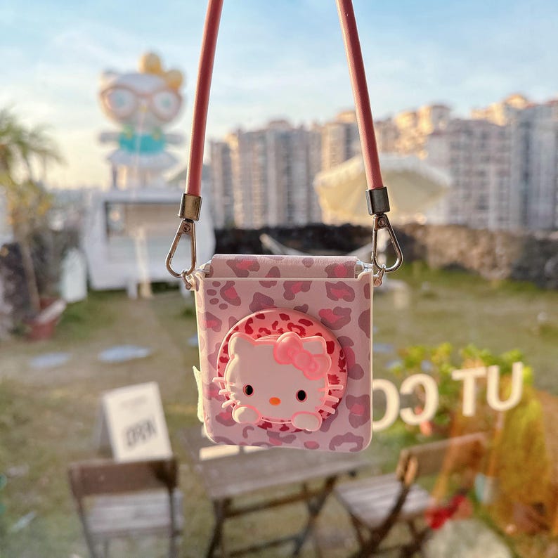 May include: Pink crossbody phone case featuring a Hello Kitty design. The case has a leopard print pattern and a 3D Hello Kitty face with a pink bow. It includes a pink strap and silver-colored metal clasps.