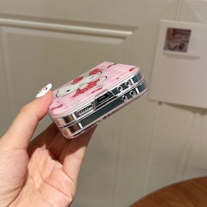May include: A pink phone case featuring a Hello Kitty design with strawberries and a gingham pattern. The case has a clear, protective border and a silver-colored frame. The design is a cute and playful accessory.