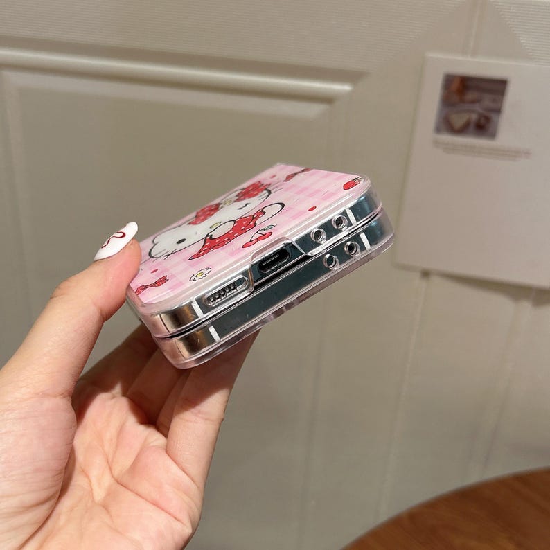 May include: A pink phone case featuring a Hello Kitty design with strawberries and a gingham pattern. The case has a clear, protective border and a silver-colored frame. The design is a cute and playful accessory.