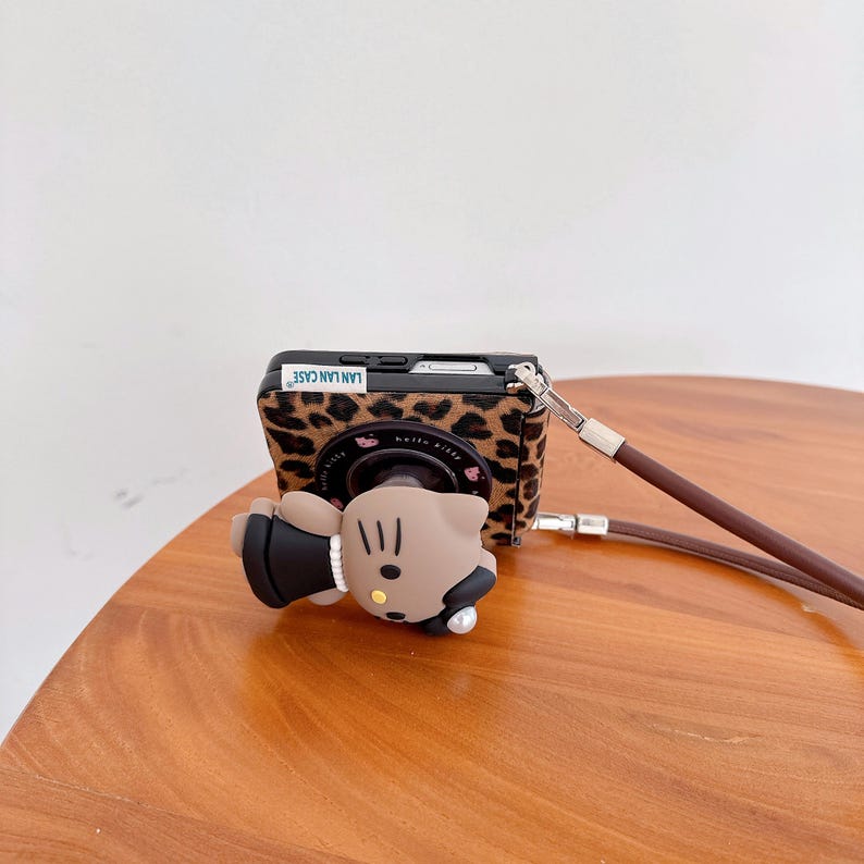 May include: A phone case with a leopard print design and a Hello Kitty character attached. The case has a brown leather strap. The text "LAN LAN CASE" is visible on the top of the case.