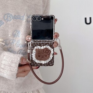 May include: A brown floral patterned phone case with a brown Hello Kitty appliqu&eacute; and a white lace trim. The case has a brown wrist strap and a silver-coloured clasp. The phone screen is black.